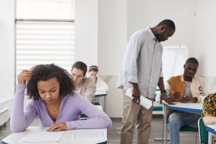 Grupo de estudantes realiza uma prova em sala de aula enquanto é supervisionado por uma professora.